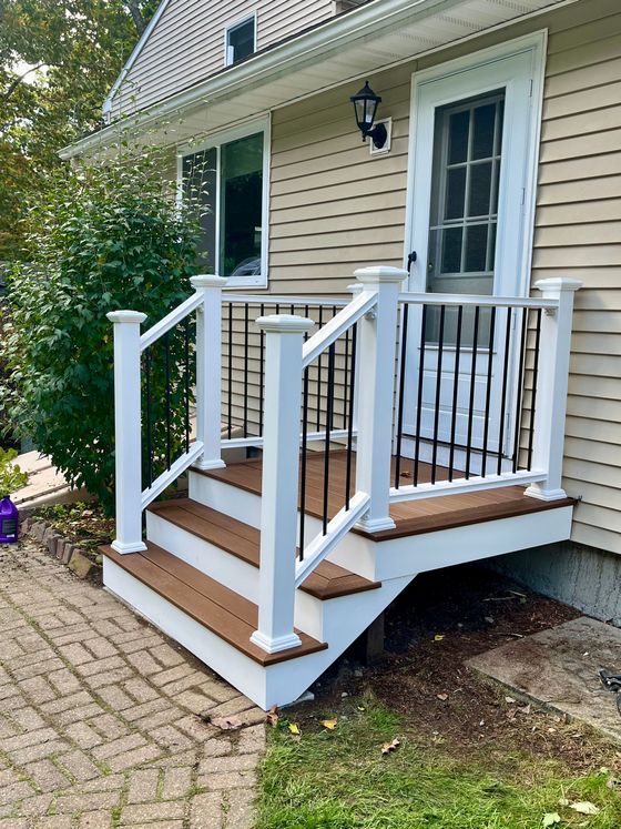 A white-railed, brown wooden front deck and stairs leading to a doorway on a house with beige siding.