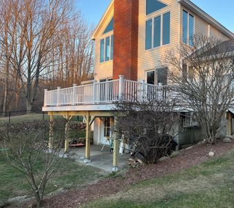 A multi-story house with light siding and a prominent stone chimney featuring a raised wooden deck with white railings.