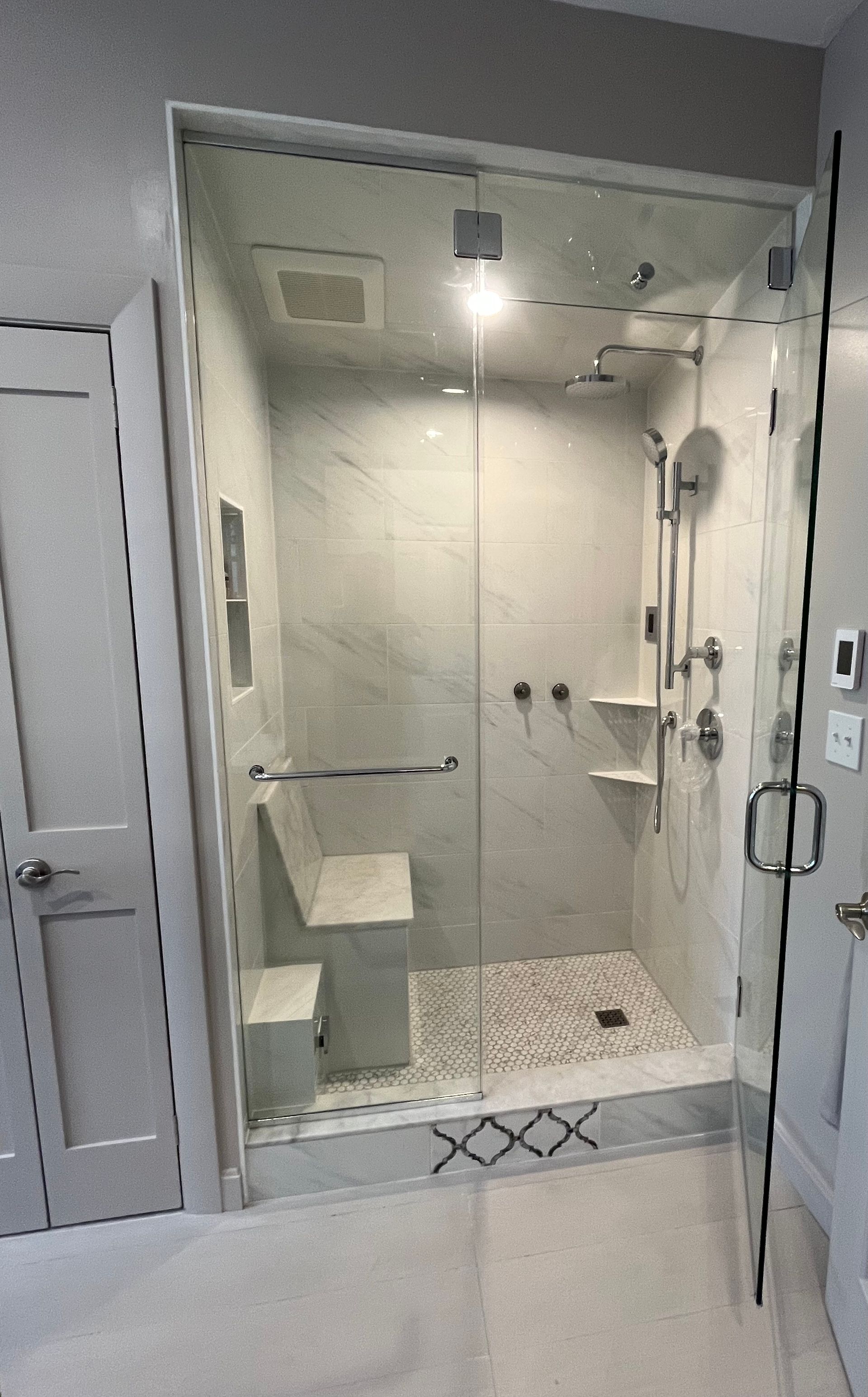 A glass-enclosed shower with white marble walls, a built-in bench, a handheld showerhead, and a decorative tile floor.