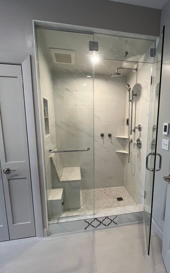 A glass-enclosed shower with white marble walls, a built-in bench, a handheld showerhead, and a decorative tile floor.