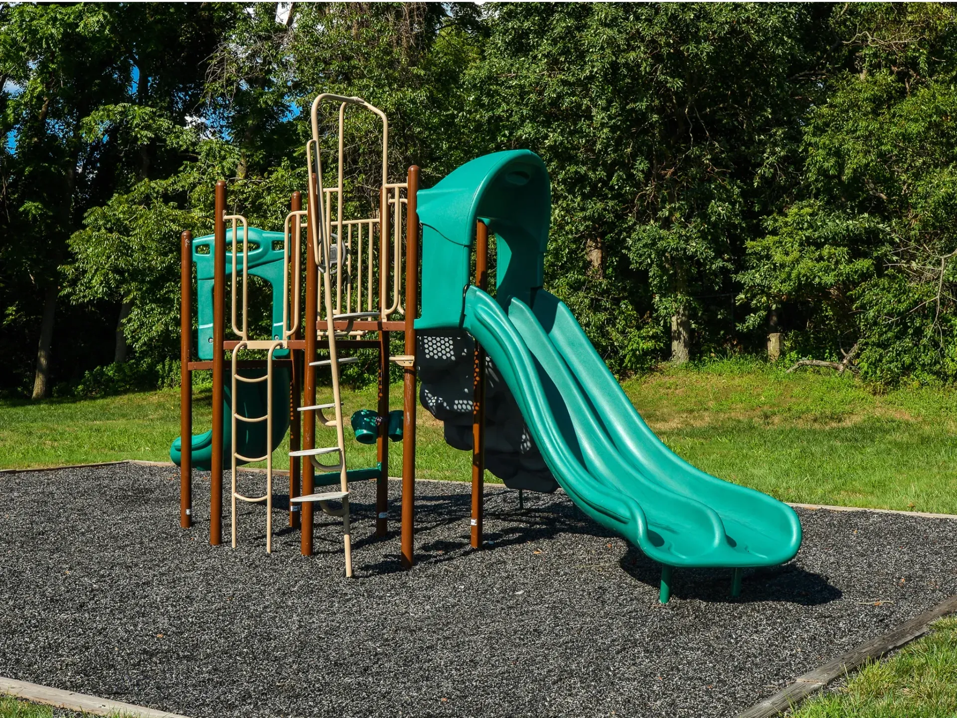 Playground with green slides and climbing structure on rubber mulch, surrounded by grass and trees.