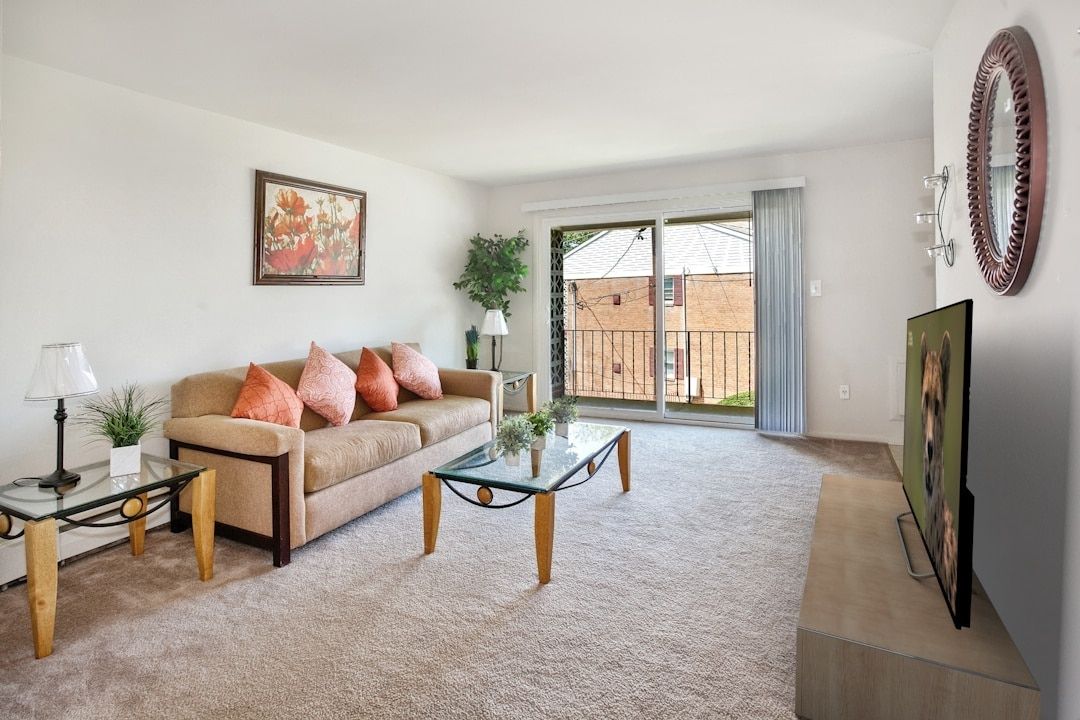 Living room in an apartment with a beige sofa, glass coffee table, TV, and sliding balcony door.