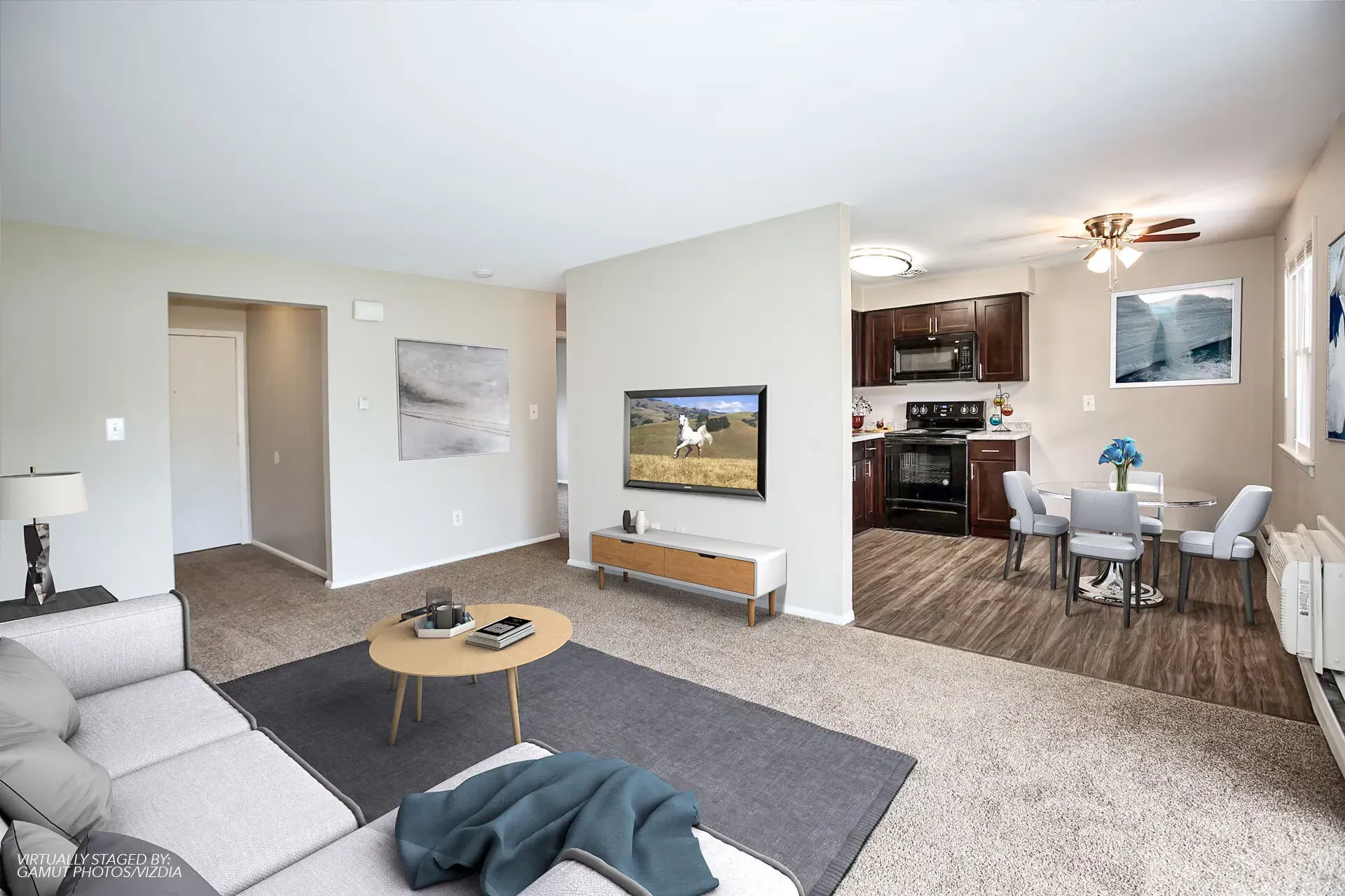 Open-concept living area with a gray sofa, wall-mounted TV, and an adjacent kitchen/dining space.