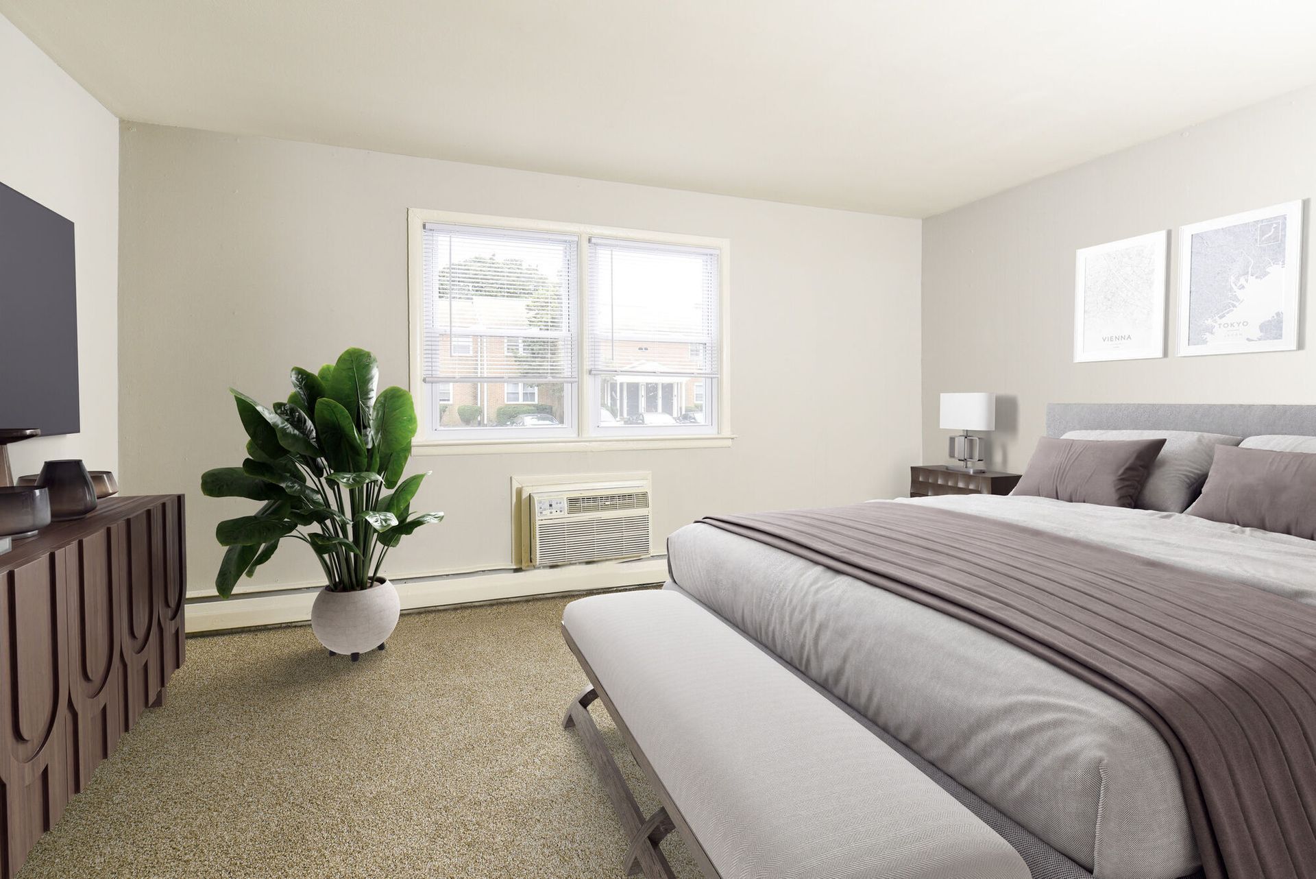 Neutral-toned bedroom in an apartment with a large bed, bench at the foot, dresser, plant, and window air conditioner.