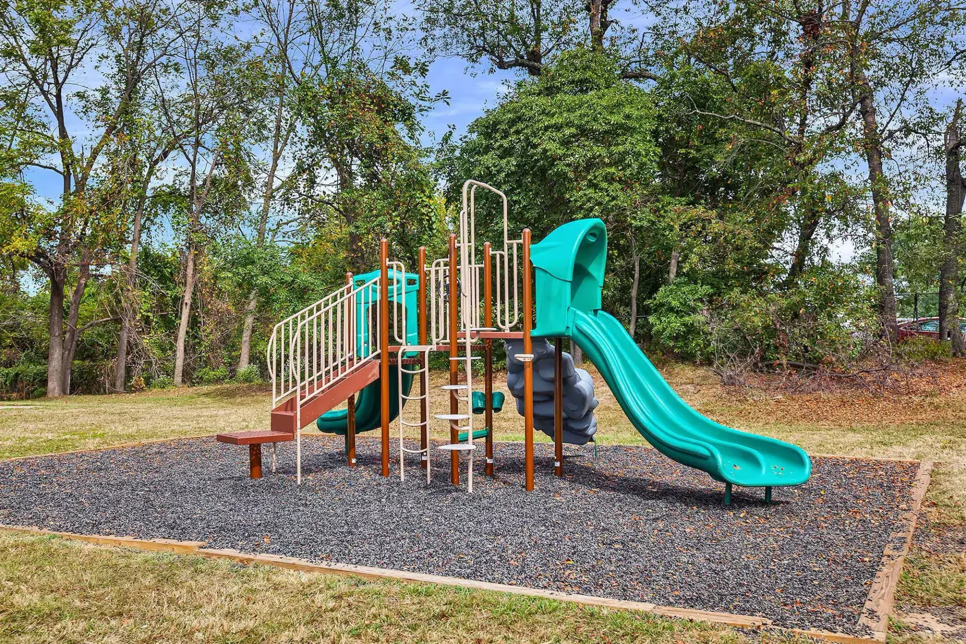 Outdoor playground with slides and climbing equipment on mulch, surrounded by trees.