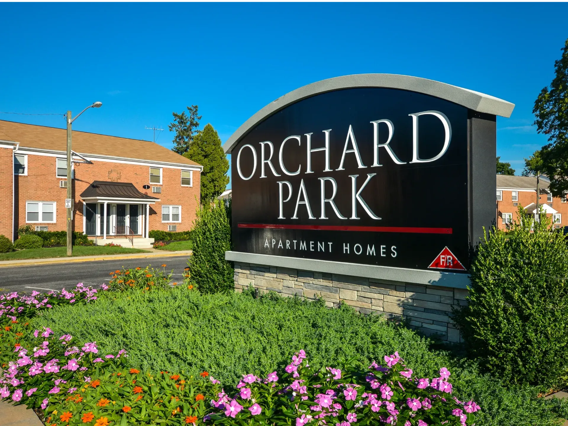 Front sign reading 'Orchard Park' with brick apartment buildings and colorful landscaping.