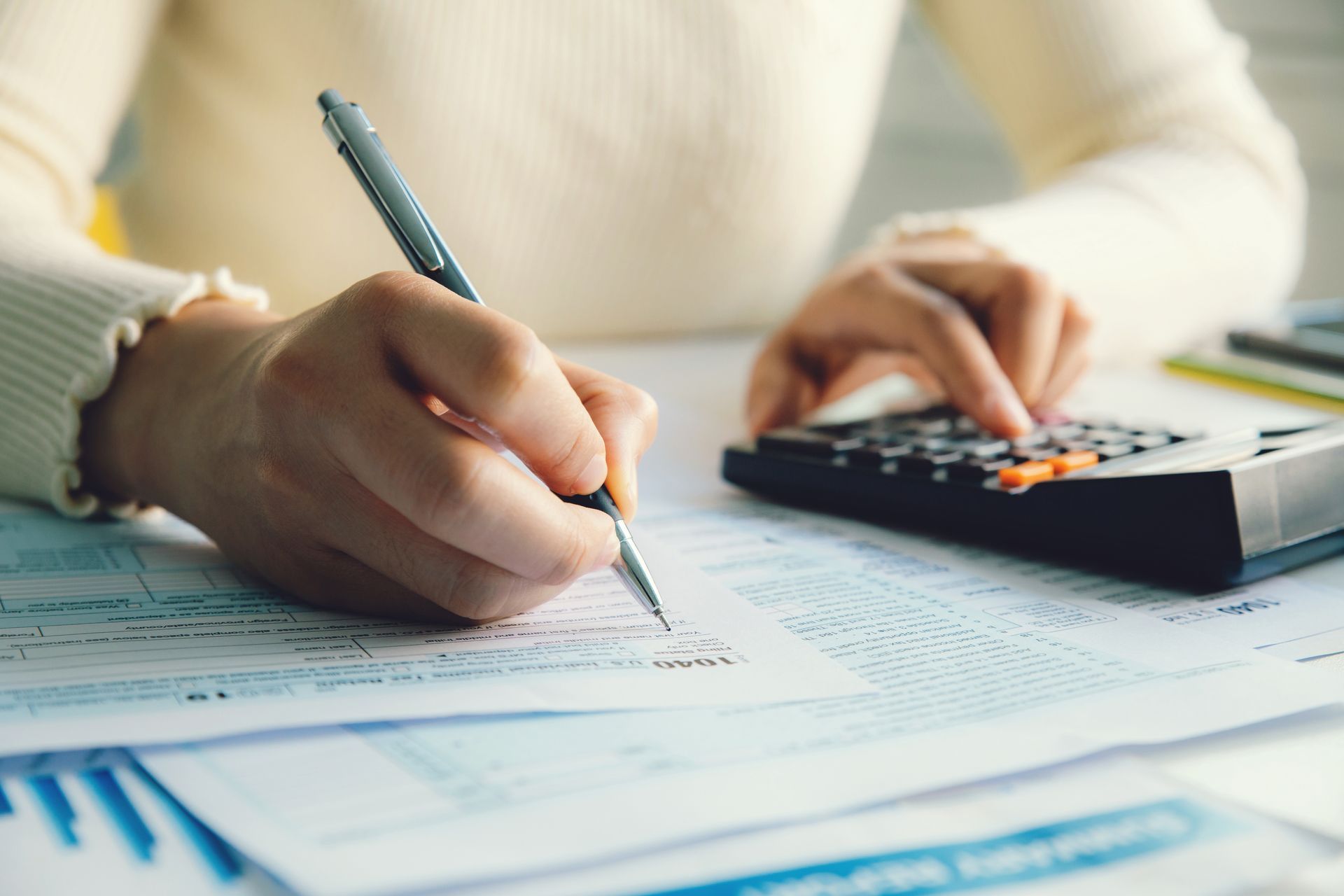 Hands filling out tax forms with a pen while using a calculator on a desk with paperwork.