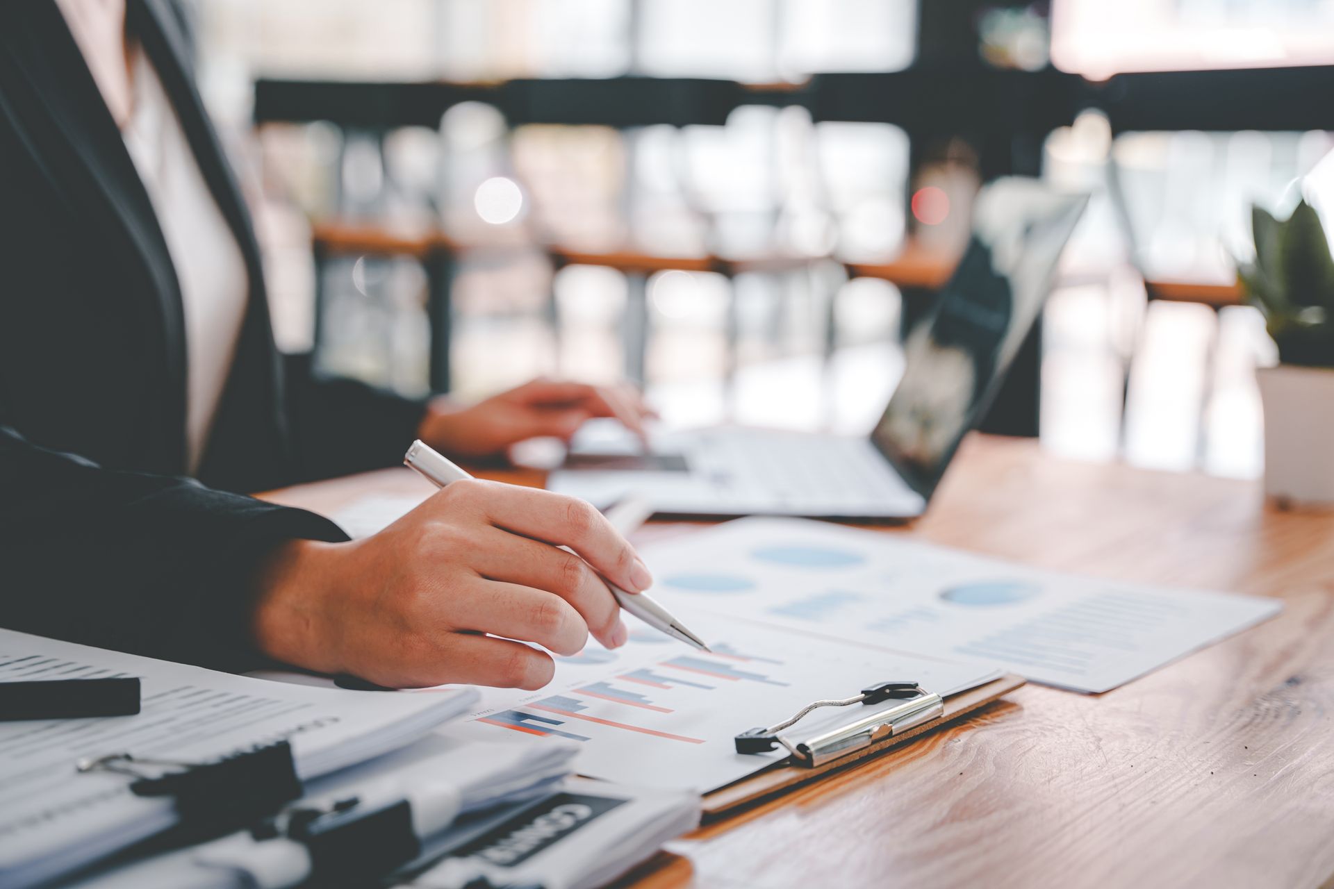 Hand holding a pen over charts on a clipboard at a desk with a laptop and documents.