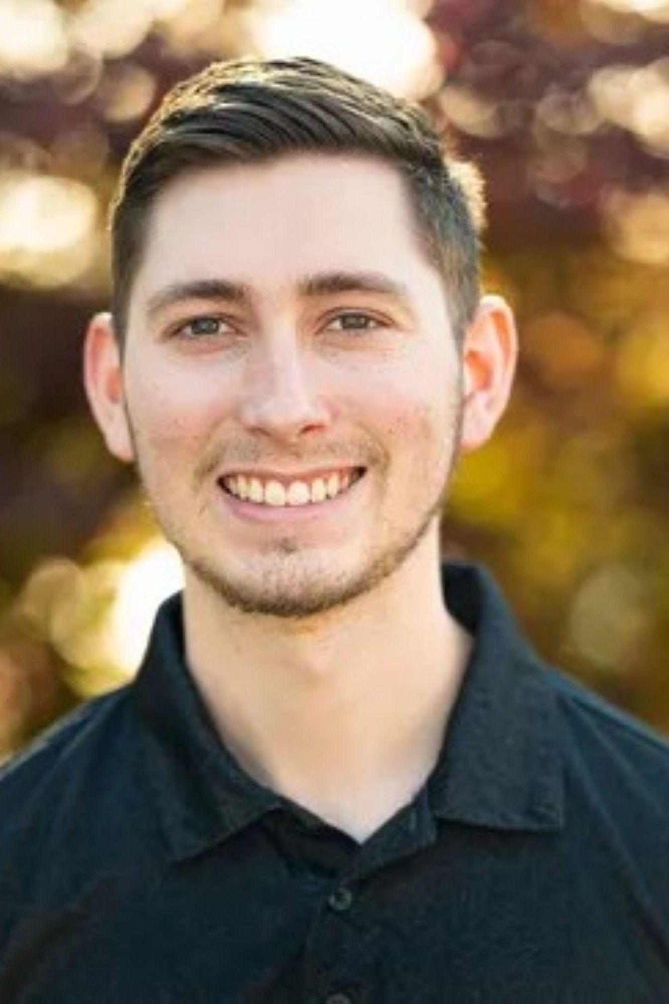Man with short brown hair smiles at the camera, wearing a dark shirt, blurred outdoor background.