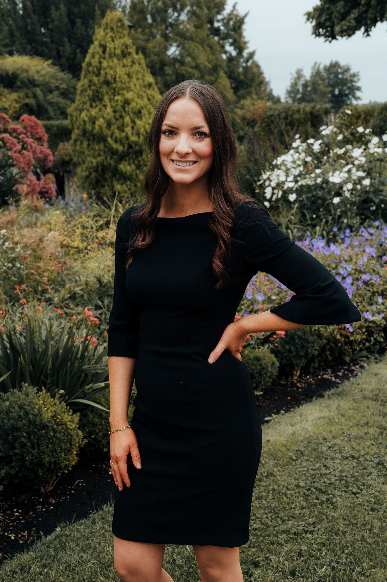 Woman in black dress poses in garden, smiling, hand on hip. Green foliage and flowers surround her.