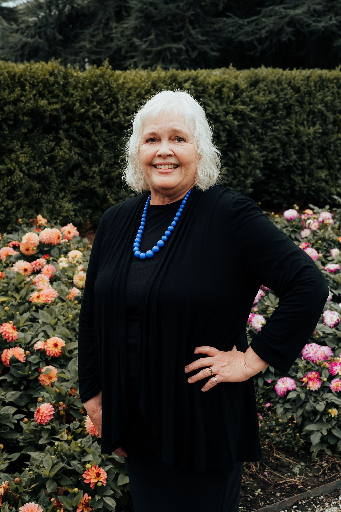 Woman in black cardigan and blue necklace poses outdoors in a flower garden, smiling.