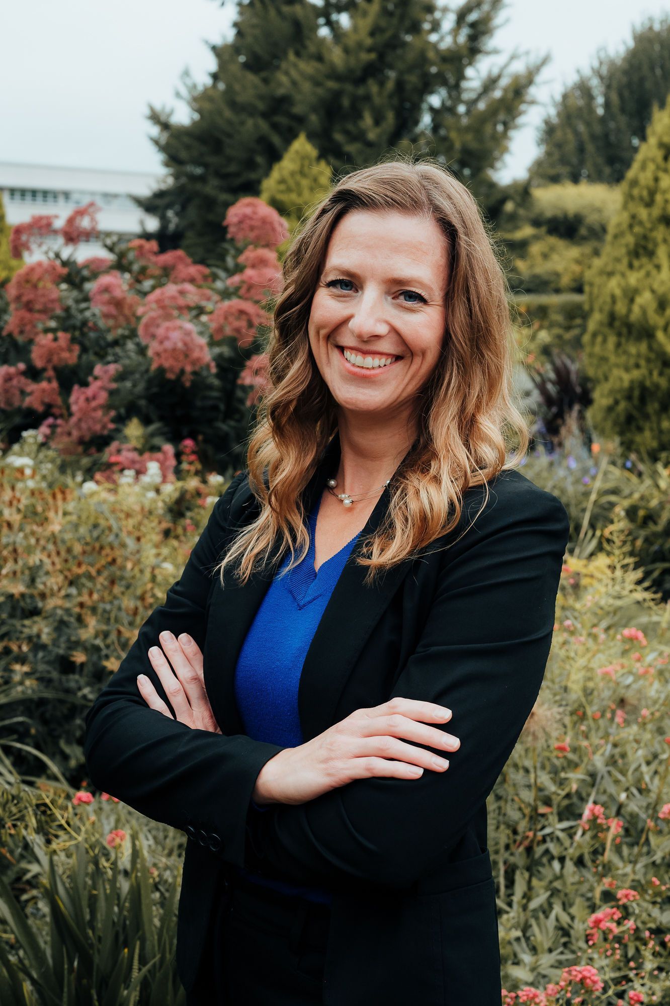 Woman in a black blazer and blue shirt smiles, arms crossed, standing outdoors among foliage.