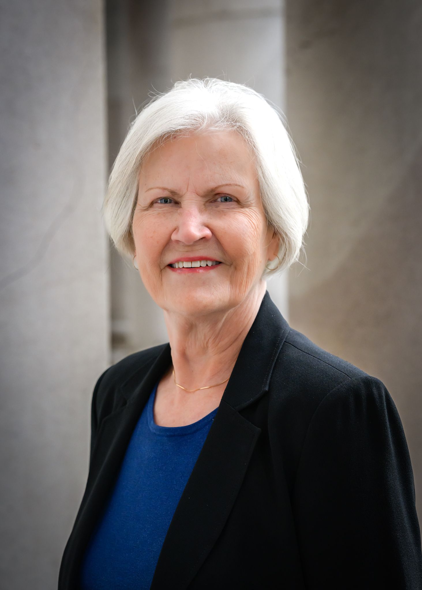 Woman with white hair in a black blazer smiles outside; columns in background.