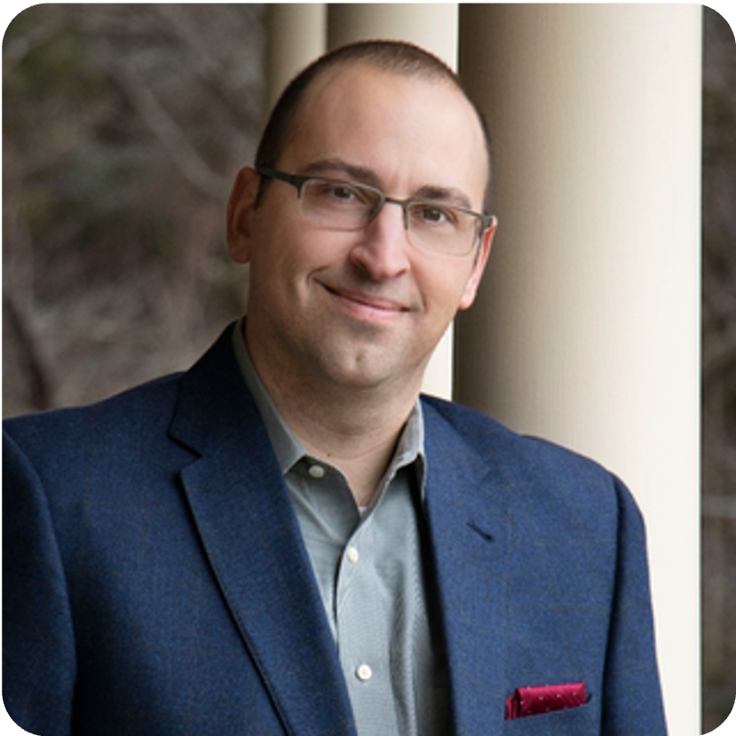 Man with glasses in a blue blazer smiles at the camera, leaning against a beige pillar in an outdoor setting.