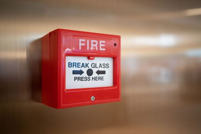 Red fire alarm box labeled “BREAK GLASS, PRESS HERE” on a blurred wall.