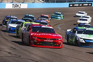 Stock cars racing through a turn, with a red Chevrolet leading a pack on the track.