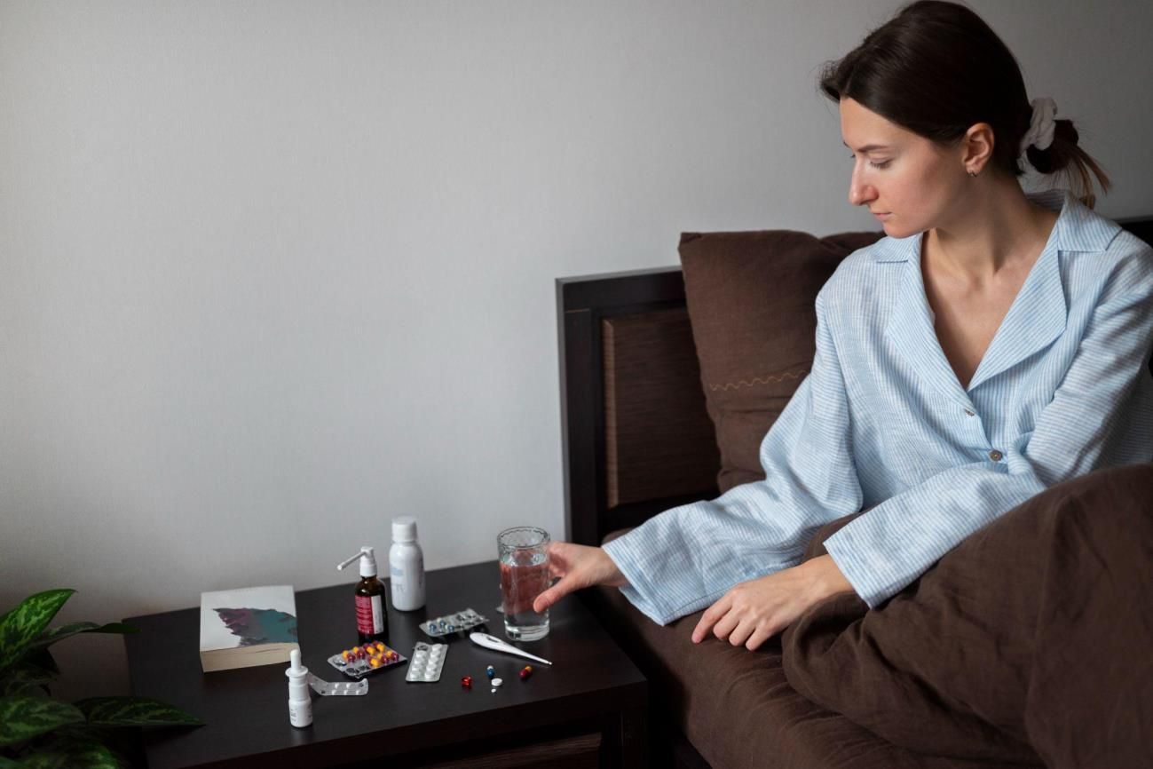 A person in light pajamas reaches for a glass of water on a side table filled with various medications and a thermometer.