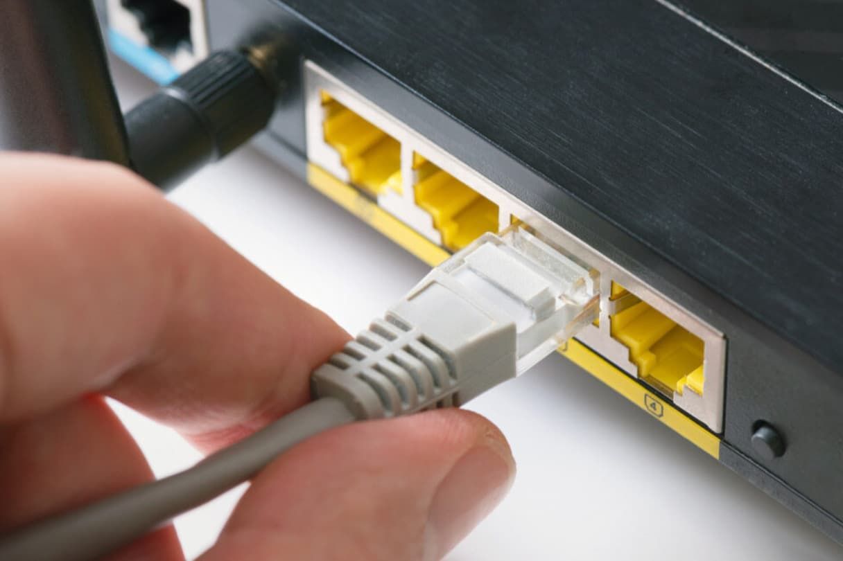 Hand Plugging A Gray Ethernet Cable Into The Yellow Port Of A Black Router — Fry Electrical In Gorokan, NSW