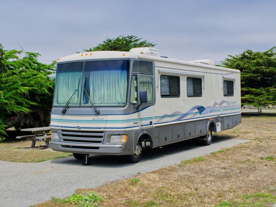 A beige and grey recreational vehicle parked on a gravel site next to green trees under a partly cloudy sky.