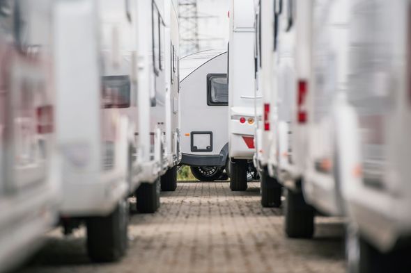 Multiple white campers parked in straight rows on a paved lot, viewed from a low-angle perspective between two vehicles.
