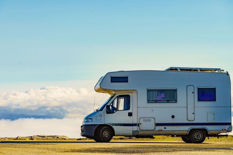 A white camper van parked on a high, rocky overlook above a layer of soft white clouds against a blue sky.