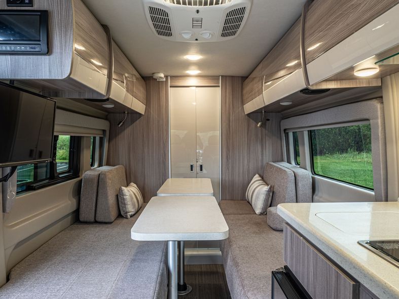 Interior view of an RV cabin with benches facing a central dining table, wood cabinetry, and a bright white closet door.