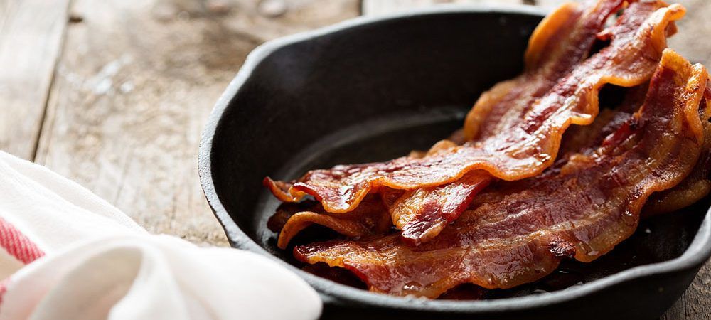 Bacon is being cooked in a skillet on a wooden table.