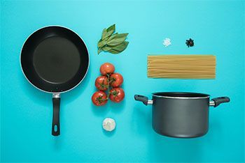 A pan , pot , tomatoes , pasta , garlic and basil on a blue background.