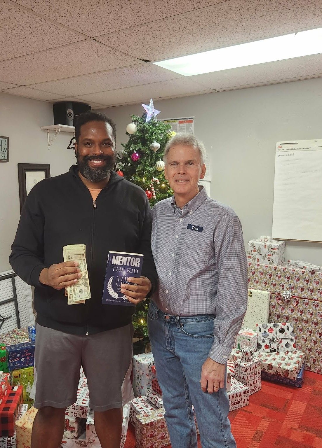 Two men stand by Christmas tree, one holds cash and a book. Presents are stacked nearby.