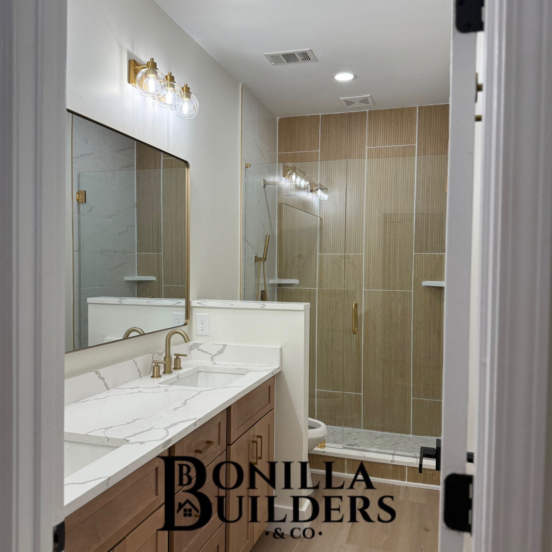 Bathroom renovation with gold fixtures, a wooden vanity, and tiled shower.