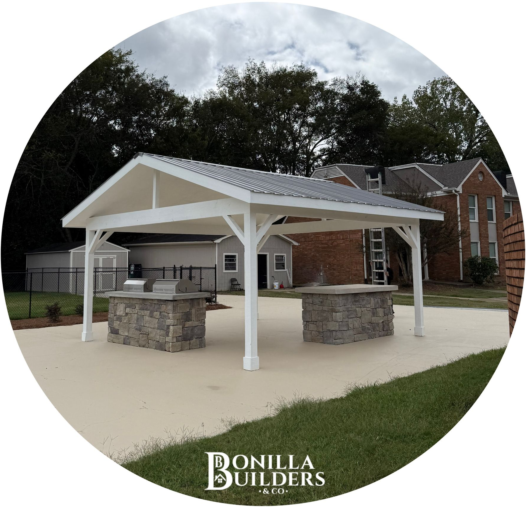 White pavilion with stone grilling stations on a concrete patio; grassy yard and trees in the background.