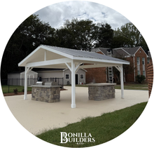White pavilion with stone grilling stations on a concrete patio; grassy yard and trees in the background.