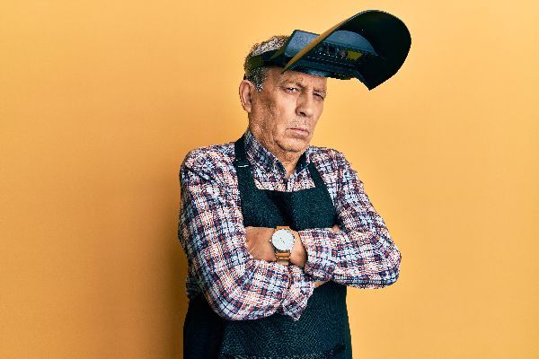 A person wearing a plaid shirt, apron, and welding helmet pushed back on their forehead, standing with crossed arms.