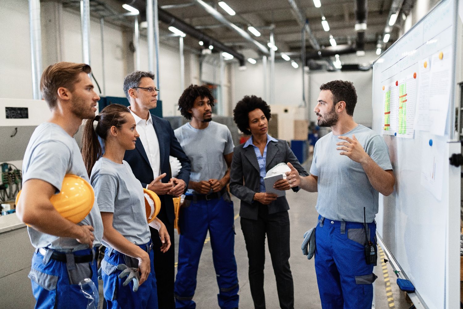 A group of workers in safety gear and two professionals in suits stand in a warehouse discussing a plan on a whiteboard.