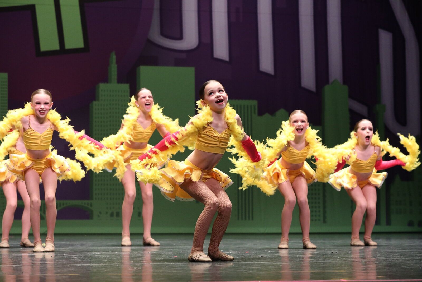 Dancers in yellow costumes with feather boas performing on stage with a city backdrop.