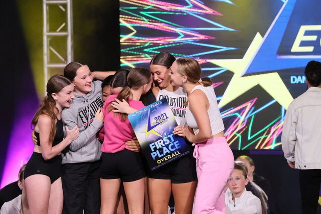Group of dancers hugging and celebrating, holding a "First Place" award on stage.