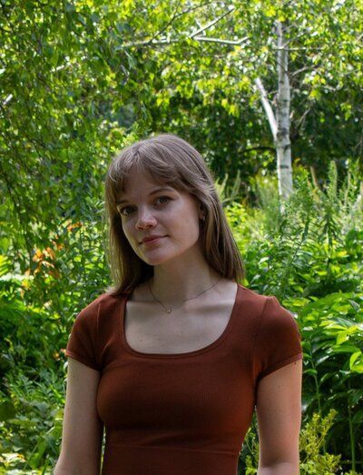 Woman with long brown hair, wearing a brown top, stands in a lush green outdoor setting.