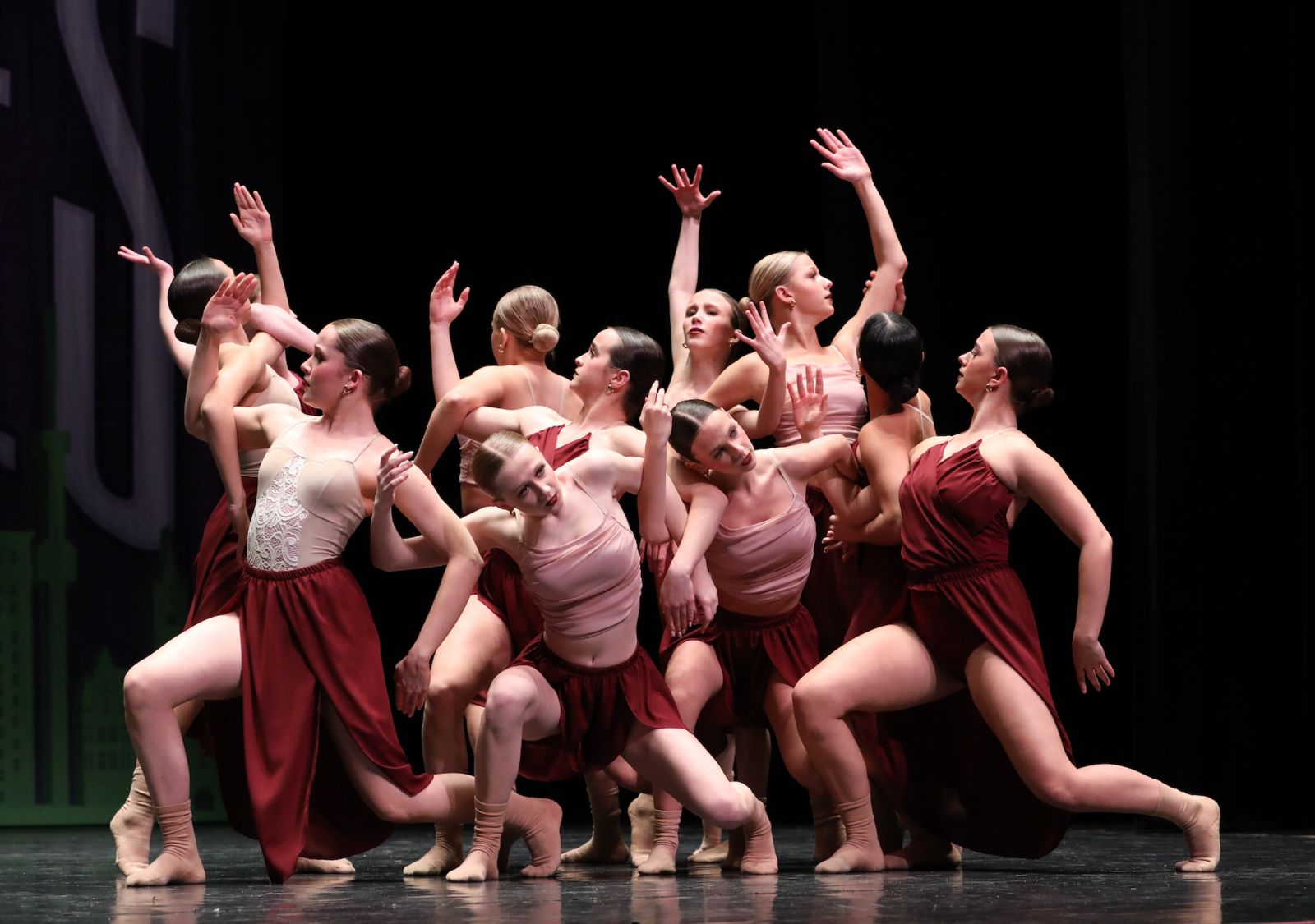 Dancers in burgundy skirts and light tops perform on stage, arms raised. Black background, stage lights.