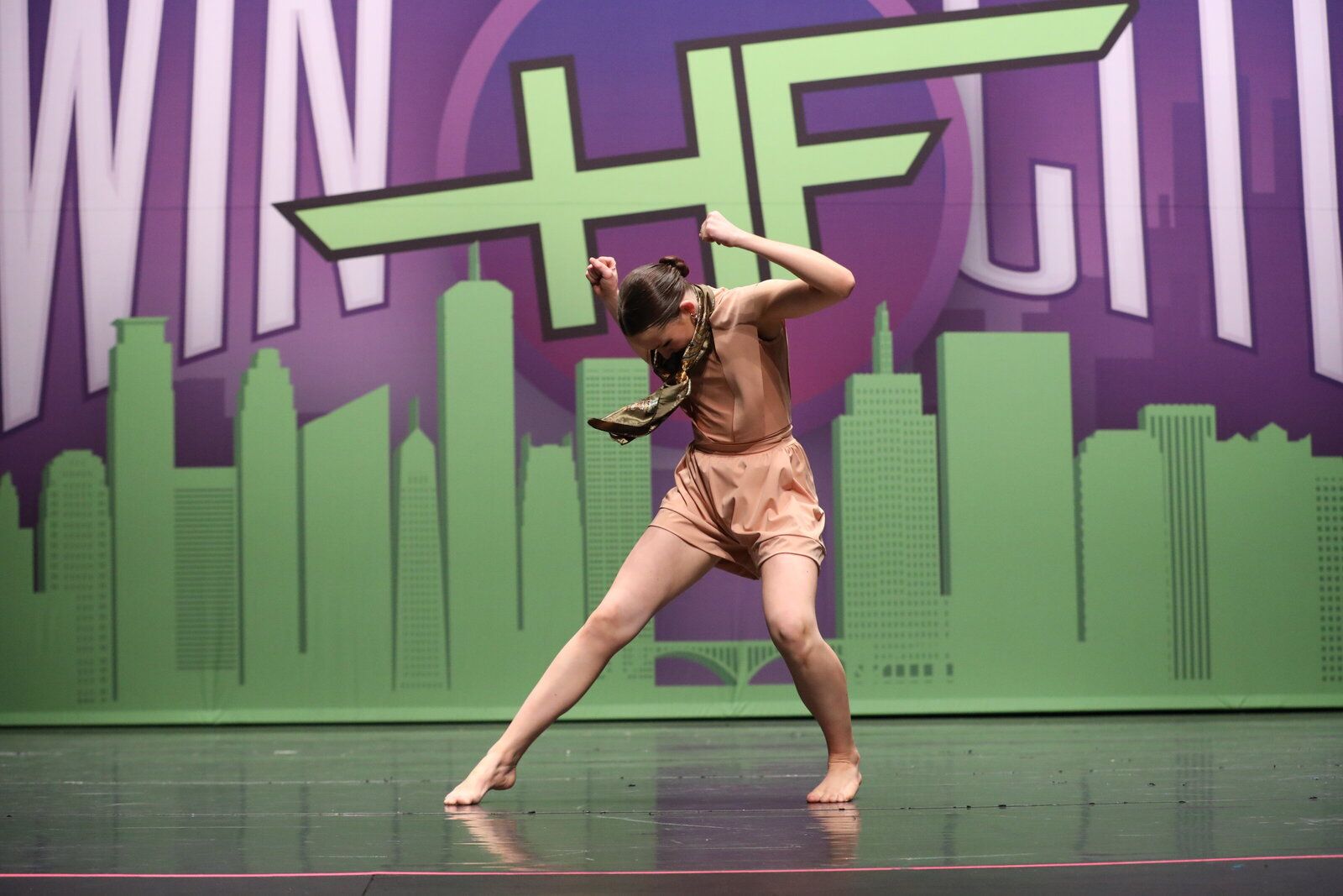Dancer in tan outfit performing on stage, bending forward with arms raised. Green background with city silhouette.