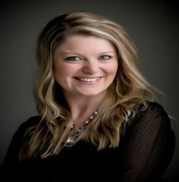 Blonde woman smiles at the camera, wearing a black top and silver necklace; studio portrait.