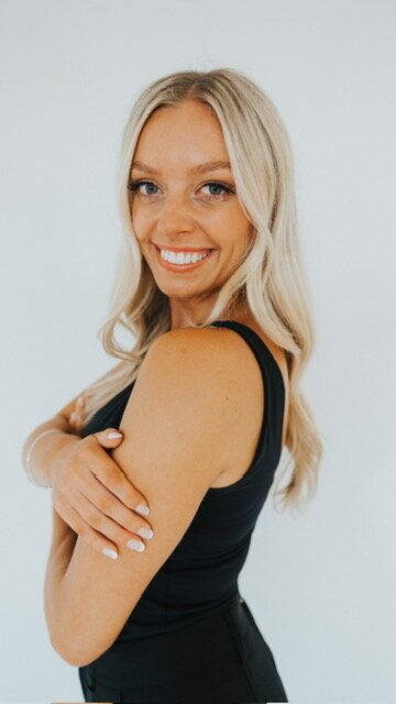 Woman with blonde hair smiles, arms crossed, in a black top against a white backdrop.