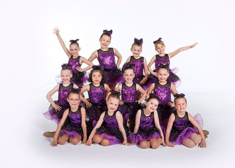 Group of dancers in matching purple and black sequined costumes posing on a white backdrop.
