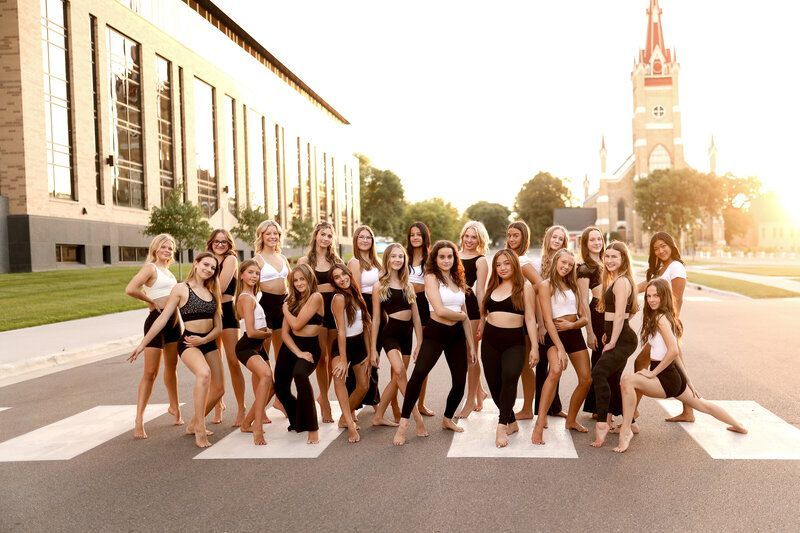 Dancers in black and white attire pose on a crosswalk; a church and building are in the background.