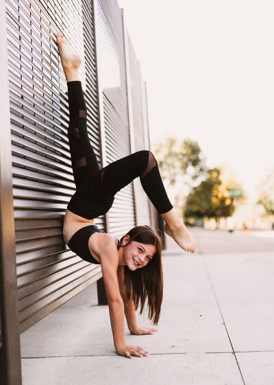 Young person in a handstand against a slatted wall, smiling. Black outfit, barefoot. Outdoor setting.