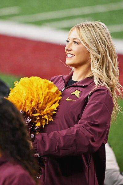 Cheerleader smiling, holding a gold pompom, wearing maroon jacket with a gold 