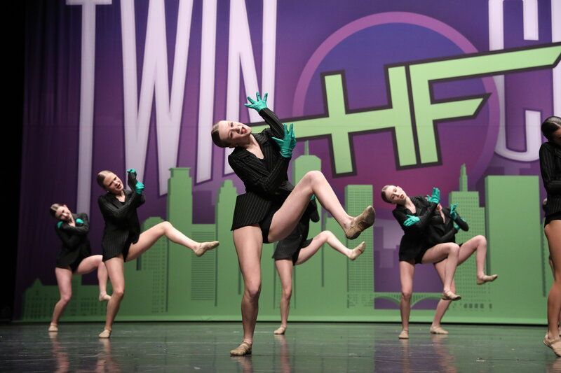 Dancers in black outfits and green gloves on stage, performing a choreographed routine.