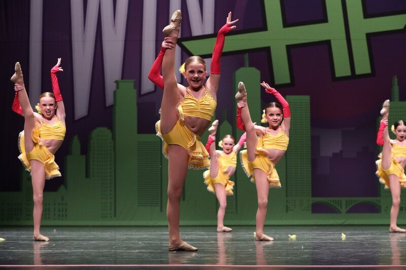 Dancers in yellow costumes perform a high kick on a stage with a cityscape backdrop; red gloves.