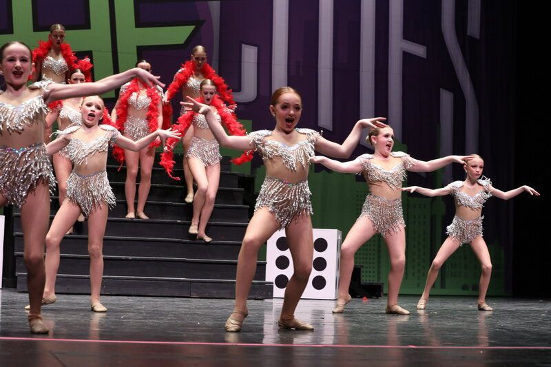 Dancers in jeweled costumes perform on stage; some on stairs, all with arms outstretched, smiles, and dice prop.