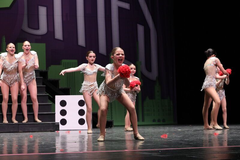 Dancers on stage, holding red pom-poms, performing in sparkly costumes. A large die prop sits nearby.