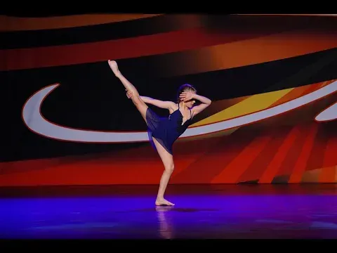 Dancer in blue leotard performs on stage, leg extended high, arms overhead. Colorful backdrop.